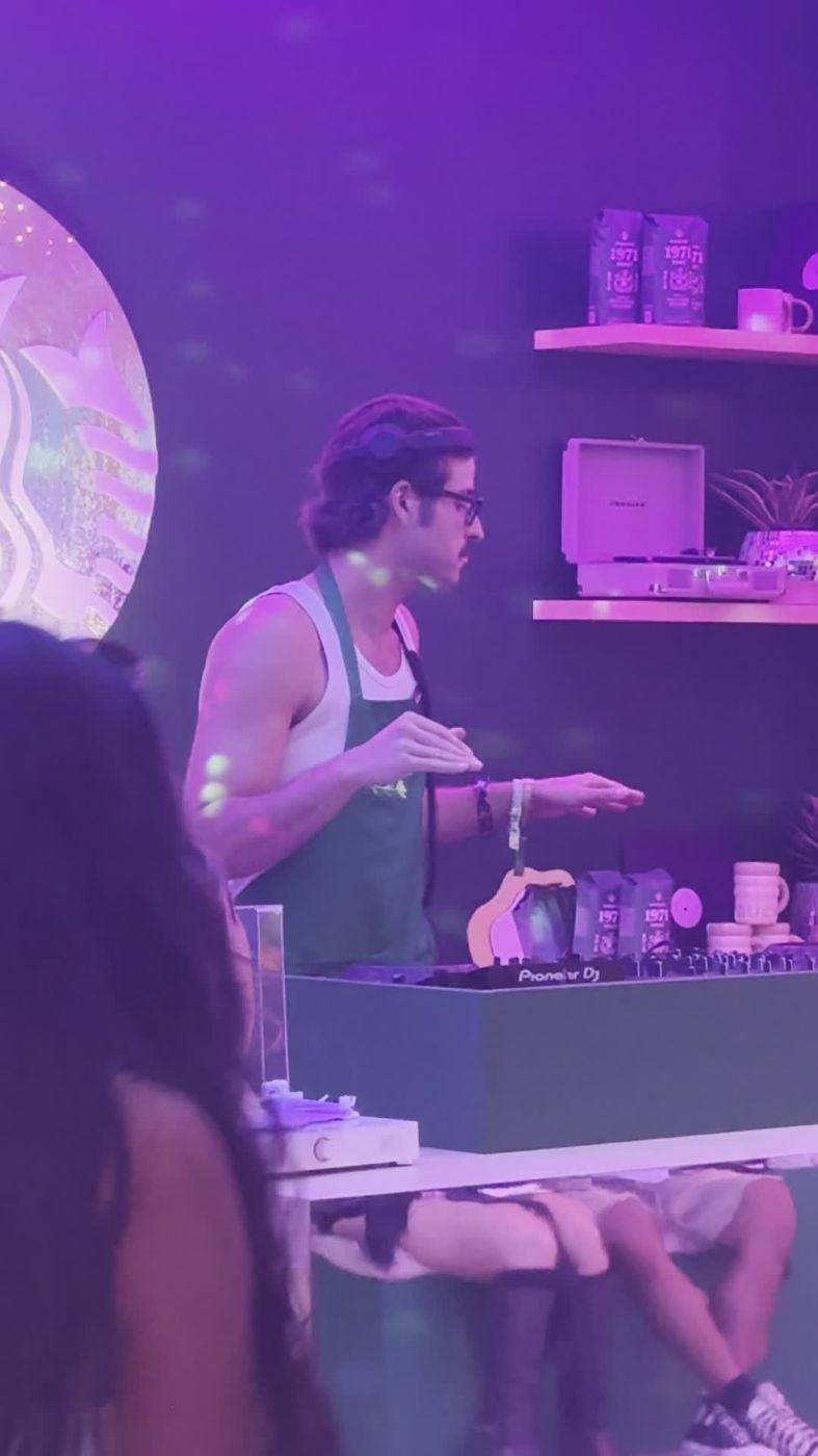 A DJ in a green Starbucks apron playing on Pioneer DJ decks in front of a lit Starbucks siren logo, with 1971 coffee bags and mugs on the shelves